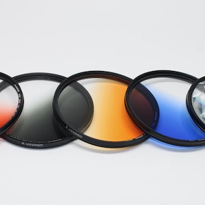 May include: A selection of camera lens filters with black frames. The filters show various colours, such as orange, black, yellow, blue, and a clear, textured filter. The filters are arranged in a line against a white backdrop.