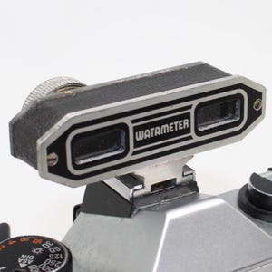 May include: A black and silver Watameter rangefinder accessory mounted on a silver camera. The device features a rectangular body with a textured black finish, a silver metal frame, and two glass viewports with the brand name printed in white.