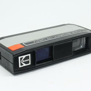 以下が含まれることがあります： ヴィンテージのKodak Instamatic 130カメラ。カメラは長方形で、黒いボディ、グレーのトップ、オレンジ色のアクセントがあります。前面にはKodakのロゴが見えます。カメラにはレンズとビューファインダーが付いています。