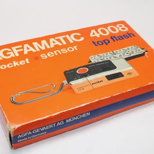 Op de afbeelding: Een oranje kartonnen doos voor een Agfamatic 4008 pocket sensor camera met flitser. De doos bevat witte en blauwe tekst, waaronder de merknaam, het model en de tekst Made in Germany. Een zilverkleurig polsbandje is aan de camera bevestigd.