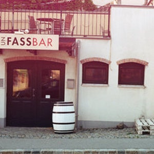 May include: A white building with a black door and a sign that says "Fassbar". There are two wooden barrels in front of the door.