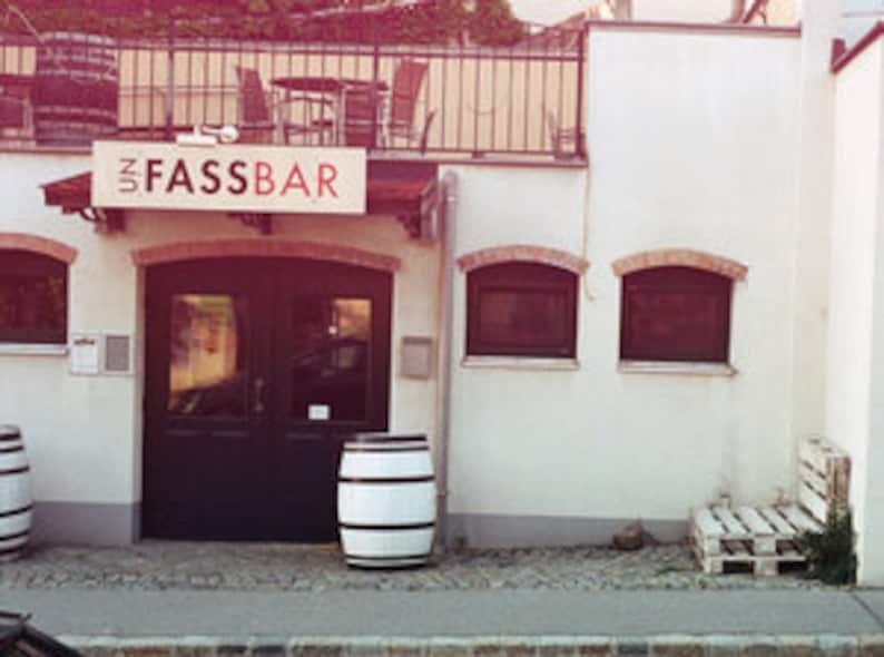 May include: A white building with a black door and a sign that says "Fassbar". There are two wooden barrels in front of the door.