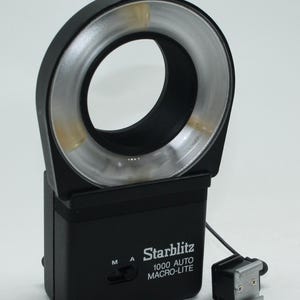 Könnte beinhalten: Ein schwarzer Starblitz 1000 Auto Macro-Lite Ringblitz für Makrofotografie. Der Blitz hat ein kreisförmiges Licht mit einem silbernen Reflektor und einem schwarzen Gehäuse. Er enthält einen Blitzschuh und ein Anschlusskabel.