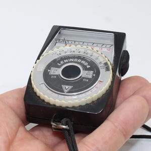 Può includere: Un esposimetro Leningrad 4 nero e argento con quadrante circolare con scale ASA e DIN, un ago indicatore e una ghiera di regolazione bianca dentellata. Il dispositivo ha un corpo in plastica nera e un cordino nero fissato nella parte inferiore.