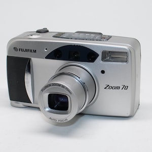 May include: Silver Fujifilm Zoom 70 digital camera with a black grip. The camera has a zoom lens and a built-in flash. The lens is labeled Fujinon Zoom 35-70mm and Auto Focus. The camera is on a white background.
