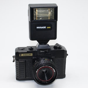 May include: A black vintage camera with a flash attachment. The camera has a lens with the text "NEW COLOR OPTICAL LENS 50mm 1:6". The flash is labeled "MIRAGE 380". The camera body is marked "BARCLAYS".