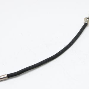 Puede incluir: Cable disparador de cámara negro con punta y botón plateados. El cable es de material tejido y está curvado. El artículo está sobre un fondo blanco.