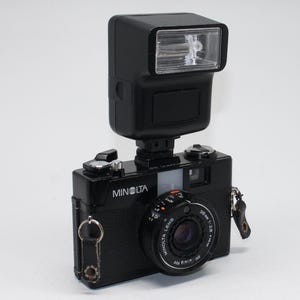 Peut inclure: Appareil photo argentique Minolta noir de 35 mm avec flash. L'appareil photo est équipé d'une lanière en cuir noir et d'un objectif "Minolta Lens 38mm 1:2.8". Le flash est monté sur le dessus.