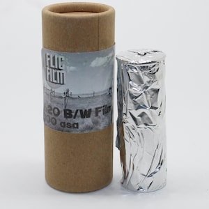 Op de afbeelding: Een rol 120 B/W film, verpakt in folie, naast een bruine kartonnen koker met een label met de tekst "FLIC FILM". Het label toont een zwart-wit landschapsfoto. De film is 100 asa.