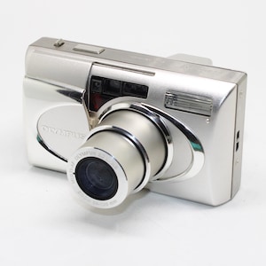 May include: A silver Olympus point-and-shoot film camera with a retractable lens. The front features the Olympus logo, a flash unit, and a lens barrel labelled Olympus ED Lens 38-105mm. The camera body has a metallic finish with a shutter button.