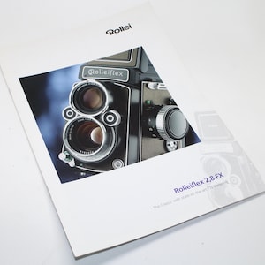 Puede incluir: Un folleto con una cámara Rolleiflex 2,8 FX. La cámara es negra y plateada con dos lentes y un dial. El folleto tiene la palabra "Rollei" en la parte superior y "Rolleiflex 2,8 FX" en la parte inferior.