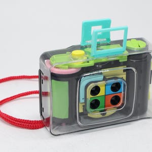 May include: A brightly coloured toy camera with a clear plastic case, featuring green, yellow, and blue accents. It has a red strap and a multi-coloured lens design. The camera is designed for children.