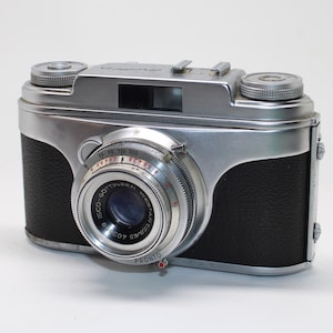 May include: A vintage silver and black film camera featuring a prominent circular lens with the text ISCO-GÖTTINGEN WESTAR 1:2.8/45 and PRONTO printed on the front. The camera body has a textured black grip and a metallic silver top plate.