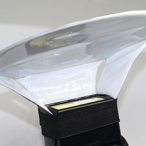 Peut inclure: Un diffuseur de flash en plastique transparent fixé à un flash d'appareil photo noir. Le diffuseur a une forme incurvée en forme d'aile et est maintenu en place par une sangle noire. Le flash a une source de lumière rectangulaire.