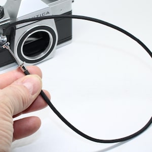 May include: A black camera shutter release cable with a silver metal tip is connected to a silver and black vintage camera. The cable is looped and held in place, ready for use. The camera has the word "Pentica" on it.