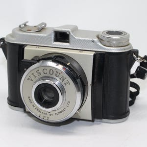 Op de afbeelding: Een zilveren en zwarte Viscount 35mm filmcamera met een zwarte leren riem. De camera heeft een lens met de tekst "Viscount Made in England by Coronet Ltd."