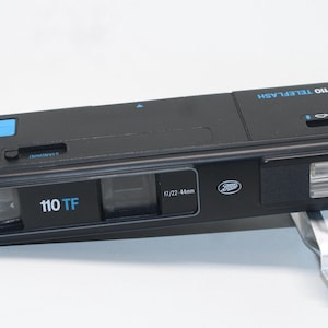 May include: Black 110 TF camera with a flash and a silver wrist strap. The camera has the text "110 TF" in blue and the Boots logo. A roll of Tiger 200/50 film is next to the camera.