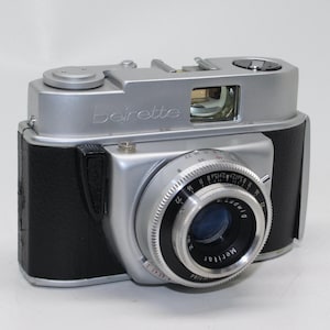 May include: A vintage silver and black Beirette camera with a Meritar lens. The camera has a silver body with black leatherette sides and a lens with a blue tint. The lens has the text "Ludwig Meritar" printed on it.