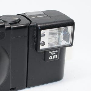 Puede incluir: Flash electrónico Olympus A11 negro. El flash tiene forma rectangular con un cabezal de flash prominente y el texto "Electronic Flash A11". La unidad también tiene el texto "ASA" y "Olympus".
