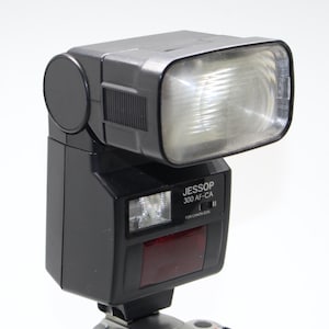 Puede incluir: Un flash de cámara negro Jessop 300 AF-CA con un cabezal de flash rectangular y un panel rojo. El flash tiene el texto "JESSOP 300 AF-CA" y "FOR CANON EOS" impreso en el lateral. Una pieza redonda negra está en el lado izquierdo del cabezal del flash.