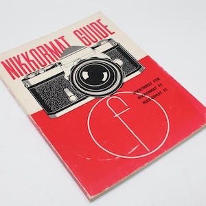 Puede incluir: Un libro guía Nikkormat vintage con una cubierta roja y crema. La portada presenta una ilustración en blanco y negro de una cámara y el texto "NIKKORMAT GUIDE". El texto adicional incluye "NIKKORMAT FTN, FT, FS".