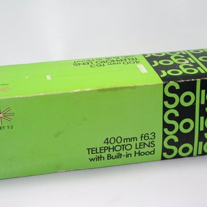 Puede incluir: Una caja verde con el texto "400mm f6.3 TELEPHOTO LENS with Built-in Hood" y la palabra "Soligor" repetida tres veces en letras negras en la parte superior y lateral de la caja. La caja tiene un logotipo de estrella blanca con el texto "PRESET T-2" en la parte inferior izquierda.