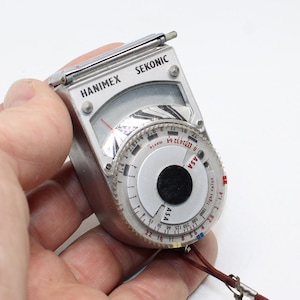 May include: A silver Hanimex Sekonic light meter featuring a circular dial with black and red text, including ASA and light value markings. The device has a needle display window at the top and a thin brown cord attached to the bottom corner.