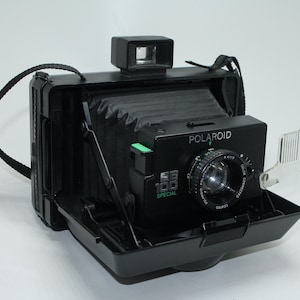 Könnte beinhalten: Schwarze Polaroid EE100 Special Sofortbildkamera mit schwarzem Riemen. Die Kamera hat einen Sucher, ein Objektiv und einen Blitzschutz. Die Kamera ist geöffnet und zeigt den Balgen und das Objektiv. Das Wort "Polaroid" ist aufgedruckt.