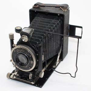 May include: A vintage black folding camera with a bellows mechanism, featuring a Voigtländer lens assembly with a Compur shutter. The device includes a metal frame viewfinder, a top-mounted handle, and silver-toned adjustment dials.