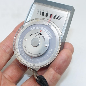 Puede incluir: Un fotómetro Boots KL-6 vintage, un dispositivo utilizado para medir los niveles de luz en fotografía. El fotómetro tiene un cuerpo plateado y gris con un dial circular y un indicador de aguja. El dial presenta números y marcas. Se adjunta un cordón negro.
