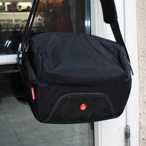 May include: Black messenger bag with a shoulder strap. The bag has a textured front panel and a red logo. The bag is hanging on a door.