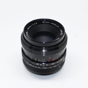 Puede incluir: Un objetivo de cámara gran angular Prinz negro con una distancia focal de 35 mm. La lente tiene marcas de apertura y enfoque, incluyendo f/5.6, f/8, f/11 y f/22. La lente está etiquetada como "WIDE-ANGLE" y "PRINZ".