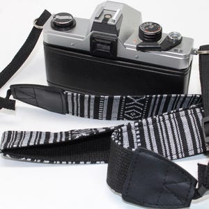 May include: A silver and black film camera with a black leather-effect body, featuring a detachable camera strap. The strap has a woven black and white geometric pattern, black nylon webbing, and black faux leather end tabs with metal hardware.