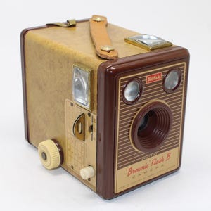 May include: A vintage Kodak Brownie Flash B box camera with a tan textured body and dark brown plastic front panel. The front features the text Kodak, Brownie Flash B Camera, and Made in England by Kodak Limited London, with two circular lenses.