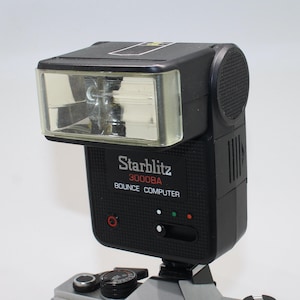 Puede incluir: Un flash Starblitz 3000BA Bounce Computer negro. El flash tiene un cabezal rectangular con una parte delantera transparente y un cuerpo negro texturizado. El texto "Starblitz 3000BA Bounce Computer" está impreso en la parte delantera.