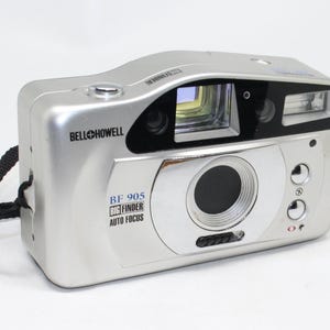 May include: A silver Bell & Howell BF 905 Big Finder Auto Focus camera. The camera has a black strap and a lens with a flash. The camera has the words "Bell & Howell" printed on the top.