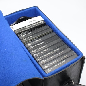 May include: A black carrying case with a blue interior holds a collection of Cokin camera filters. The filters are black with white text, including "FOG 2 A 150" and "GRADUAL Y1 A 132". The case has a handle and is open.