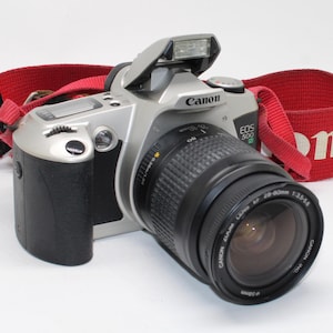 May include: A silver and black Canon EOS 500 N film camera with a 28-80mm lens and a bright red neck strap. The camera features a pop-up flash, a black rubberised grip, and clearly visible branding on the body and the lens barrel.