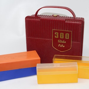Peut inclure: Une mallette rouge en plastique pour diapositives avec une poignée et un écusson doré indiquant "300 Slide File". Il y a aussi plusieurs petites boîtes de rangement de diapositives en plastique orange, bleu et jaune.