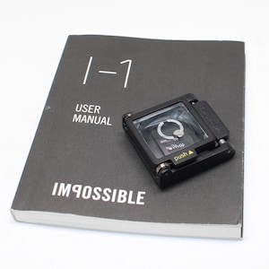 Puede incluir: Un manual de usuario negro de Impossible I-1 con texto en blanco y un pequeño accesorio de cámara cuadrado negro con lente transparente, apertura circular y una etiqueta amarilla de push sobre la cubierta.