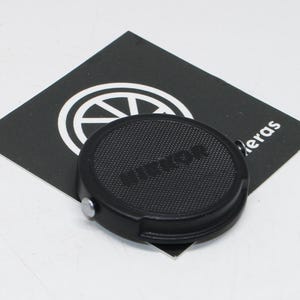 Op de afbeelding: Een zwarte Nikon lensdop met de merknaam in reliëf op de voorkant. De dop is rond met een gestructureerd oppervlak en een kleine metalen knop. Hij ligt op een zwarte kaart met een wit logo en het woord "leras".