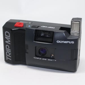 Puede incluir: Una cámara de película Olympus Trip MD 35 mm negra con flash incorporado. La cámara tiene un botón rojo en la parte superior y el texto "TRIP MD" en el lateral.