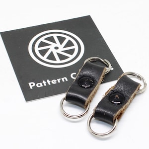 May include: Two black leather key keepers with silver-toned metal rings at each end and a central black snap button closure. A black square card with a white circular wheel icon and the text Pattern C is positioned behind the leather items.