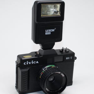 May include: Black Civica MX-V camera with a Luxon 115 M flash unit. The camera has a color lens with auto focus. The camera is black with a silver lens and a flash unit on top.