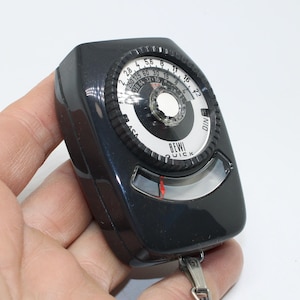 May include: A black vintage light meter, held in a hand, with a circular dial featuring numbers and the text "BEWI QUICK". The meter has a clear window and a silver metal attachment.