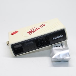 May include: A vintage Kalimar Mini 110 camera in cream with a black front panel. The camera has a red shutter button and the words "Kalimar Mini 110" in red. A silver film pack is next to the camera.
