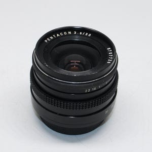 May include: A black Pentacon 3.5/30 camera lens. The lens has a series of rings with numbers and text, including "PENTACON 3.5/30" and "6707726". The lens is designed for photography and has a glass element.