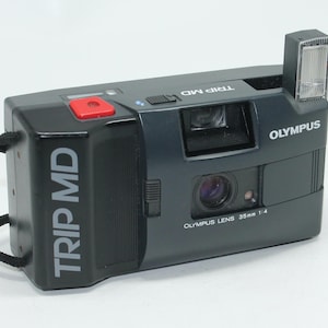 Puede incluir: Cámara de película Olympus Trip MD negra de 35 mm con flash incorporado. La cámara tiene un botón rojo y el texto "TRIP MD" y "OLYMPUS LENS 35mm 1:4" impreso. Una correa de muñeca negra está adjunta.