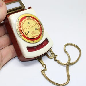 May include: Vintage Ikophot light meter in a red and cream case with a gold chain. The meter has a circular dial with numbers and the text "IKOPHOT" and "ZEISS IKON". The meter is a classic photography accessory.