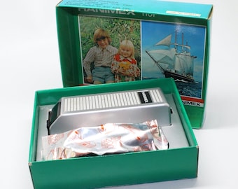Pode incluir: Uma câmera de bolso Hanimex 110 prateada na sua caixa original. A caixa é verde e tem uma imagem de um navio na frente. A câmera está na caixa com um rolo de filme de 200 ISO.